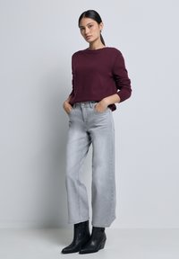 Pull bourgogne, col rond, avec des poignets côtelés ; jeans gris clair à jambes larges ; bottines noires à bout pointu ; modèle pose avec les mains dans les poches.