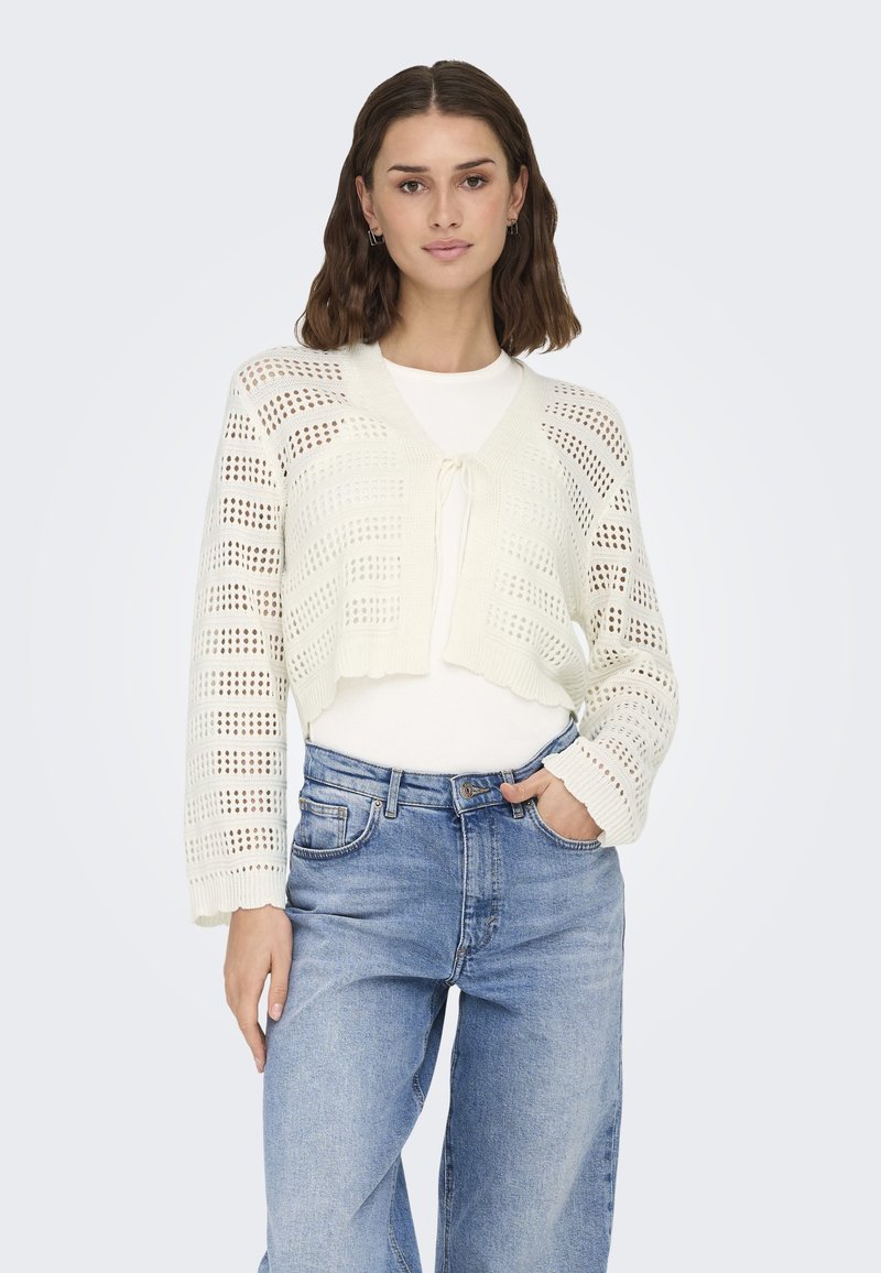 Gehaakte cardigan in off-white met open geweven patroon, strikdetail aan de voorkant en lange mouwen. Draagt over een witte top en blauwe jeans.