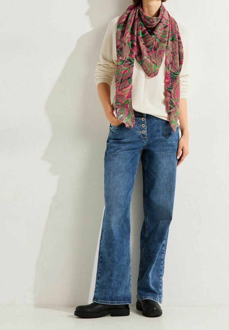 Personne portant un pull crème, un jean bleu taille haute à boutons, des bottes noires et une grande écharpe rose et verte à motifs.