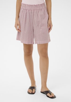 Nő rózsaszín-fehér csíkos, gumis derékpántos rövidnadrágban és fekete, nyitott orrú papucsban áll fehér padlón.