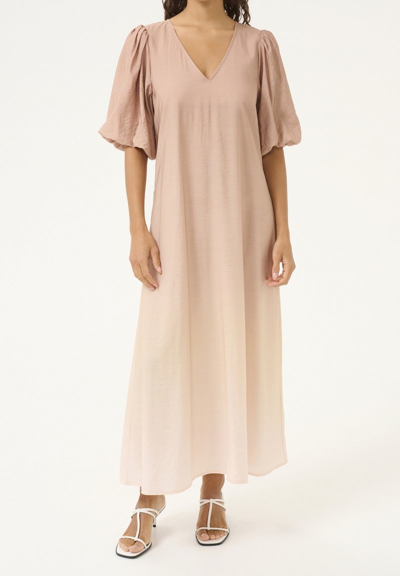 Femme portant une robe maxi rose clair à col en V avec des manches courtes bouffantes et des sandales blanches à talons avec des lanières.
