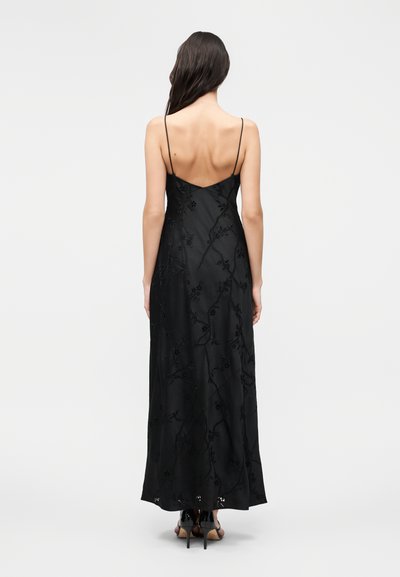 Γυναίκα με μακριά σκούρα μαλλιά που φορά μαύρο μακρύ φόρεμα με λεπτές τιράντες και λουλούδια και μαύρα ψηλοτάκουνα σανδάλια, στέκεται με την πλάτη προς την κάμερα.