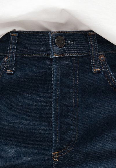 Ζώνη από σκούρο μπλε τζιν με κουμπί κλεισίματος, θηλιές για ζώνη και πορτοκαλί ραφές αντίθεσης κάτω από ένα λευκό υφασμάτινο στρώμα.