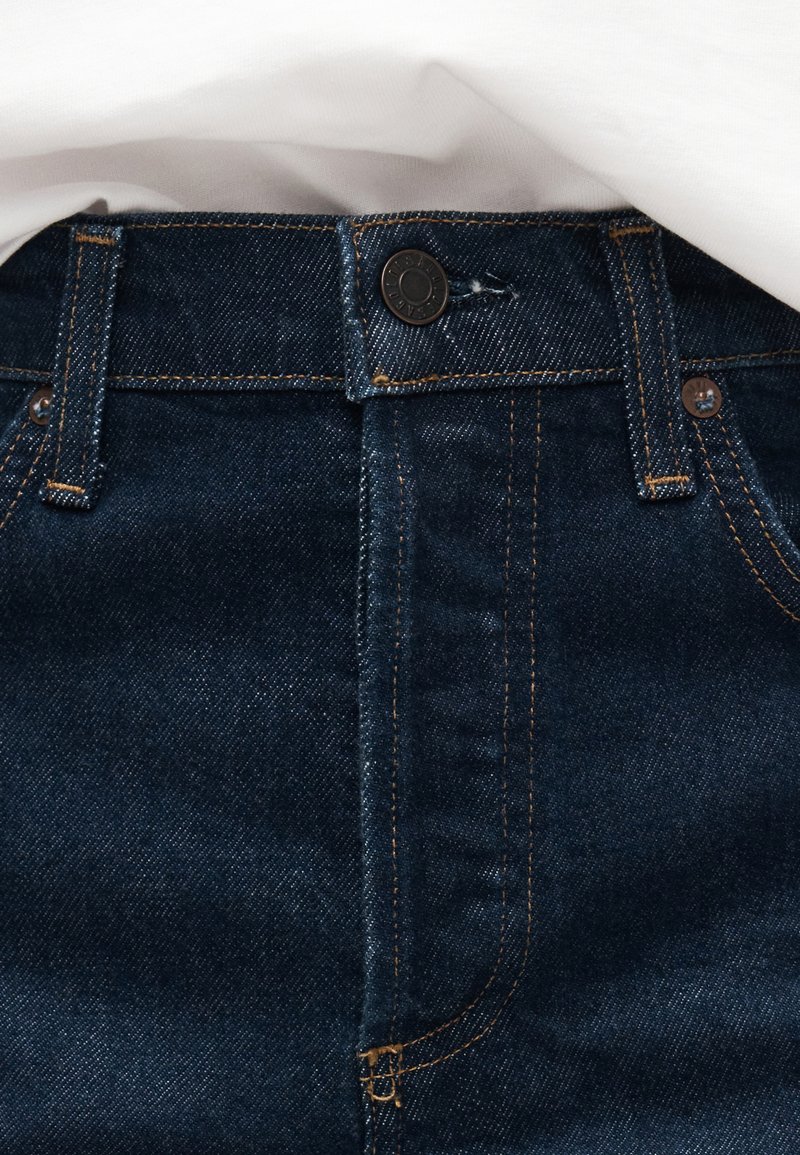 Ceinture de jeans en denim bleu foncé avec fermeture à bouton, passants pour ceinture et surpiqûres contrastantes orange sous une couche de tissu blanc.