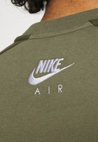 Olivově zelená mikina z látky s žebrovaným límcem, se bílým vyšitým logem "NIKE" a textem "AIR" pod ním. Detailní pohled na zadní stranu.