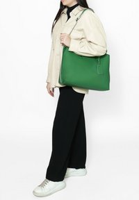 Sac à main en cuir vert au design structuré, avec une étiquette amovible et des accessoires en argent, porté à l'épaule par une personne en tenue décontractée.