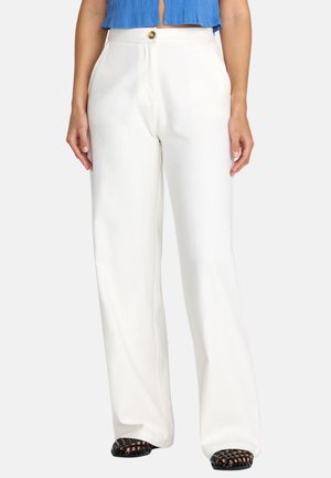 Kobieta ubrana w białe, szerokie spodnie z wysokim stanem i zapięciem na guziki, niebieski krótki top oraz czarne wiklinowe buty z odkrytymi palcami.