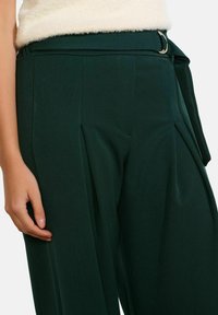 Žalios plačios kelnes su klostėmis ir audinio diržu. Medžiaga atrodo lygi, su pritaikytu liemens diržu ir šoninėmis kišenėmis.