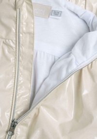 Veste beige brillante avec une fermeture éclair argentée, dotée d'une doublure en tissu blanc doux à l'intérieur. Un motif à pois est visible à l'extérieur.