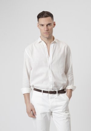 Hombre con cabello corto y oscuro, vestido con una camisa blanca abotonada con las mangas remangadas, pantalones blancos y un cinturón marrón oscuro, con las manos en los bolsillos.