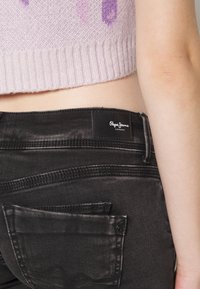 Svarta denimjeans med en bakficka med knappstängning och en märkeslapp, tillsammans med en ljusrosa kortärmad stickad tröja.