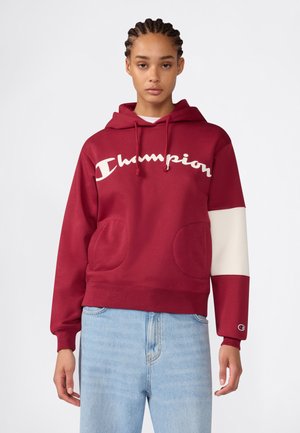 Junge Frau mit geflochtenem Haar, die einen roten Champion-Hoodie mit weißem Ärmeldetail und hellblauen Jeans vor einfachem Hintergrund trägt.
