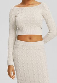 Off-the-shoulder knit top in light beige, featuring a textured heart pattern, ribbed hem, and long sleeves, paired with a matching skirt.