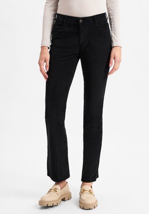 Person trägt schwarze Hosen mit geradem Bein, beige Loafers mit goldener Kettendetail und ein hellbeiges Langarmoberteil vor weißem Hintergrund.