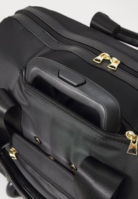 Fekete szövet táska, amely kihúzható fogantyúval, arany cipzár dísz�ítéssel és texturált felülettel rendelkezik, látható varrásokkal és fém részletekkel.