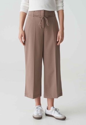 Person trägt hellbraune, weit geschnittene, gekürzte Hose mit Kordelzug in der Taille und weiße Sneakers, steht vor einfachem hellem Hintergrund.