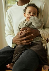 Un enfant portant un cardigan tricoté gris clair texturé avec des boutons en bois est assis sur les genoux d'un adulte ; l'adulte est habillé d'un haut blanc.