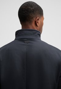 Dark gray coat with a high collar, smooth texture, and minimalistic design. Features a small logo tag at the back near the neck.