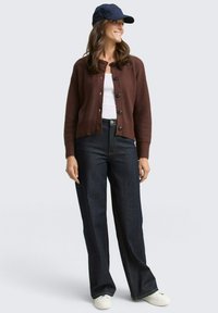 Brauner gerippter Cardigan mit schwarzen Knöpfen, getragen über einem weißen Top, kombiniert mit dunkelblauen, weiten Jeans und weißen Sneakers. Marineblaues Cap auf dem Kopf.