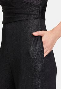 Fekete overál fényes, texturált felülettel. Kihangsúlyozott derék, oldalsó zsebek és sima, elegáns vonalvezetés jellemzi.