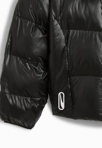 Doudoune noire avec un tissu matelassé brillant, arborant un logo blanc sur le devant et une silhouette décontractée et oversize.