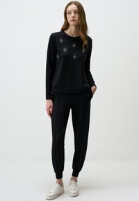 Top nero a maniche lunghe con strass decorativi, abbinato a pantaloni neri a sigaretta. L'outfit presenta una consistenza morbida e polsini elasticizzati. Le sneakers bianche completano il look.
