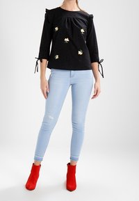 Černá blůza s volánovými rukávy a květinovou výšivkou, kombinovaná se světle modrými slim kalhotami a červenými kotníkovými botami.