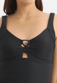 Maillot de bain noir à côtes avec une découpe en forme de goutte et un accent noué au niveau de l'encolure. Bretelles ajustables pour un meilleur ajustement. Texture lisse.