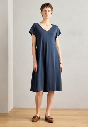 Kleid aus dunkelblauem Baumwollstoff mit kurzen Ärmeln, V-Ausschnitt, lockerer Passform und knieumspieltem Rock. Kombiniert mit braunen Slippern. Sanfte Textur.