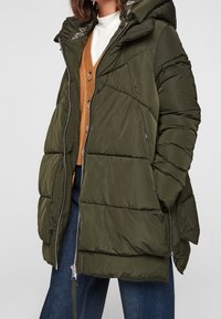 Chaqueta acolchada verde oliva con capucha, cierre de cremallera y patrón de acolchado horizontal. Combinada con un cárdigan marrón y pantalones vaqueros.