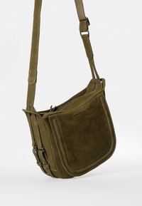 Olivgrüne Wildleder-Umhängetasche mit verstellbarem Tragegurt, Reißverschluss und zwei seitlichen Schnallen. Glatte Textur und strukturierte Form.