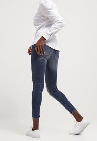 Jeans ajustados de denim azul oscuro con textura suave, que presentan un detalle desgastado en la parte superior del muslo y un ajuste ceñido, combinados con zapatillas blancas.