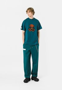 Camiseta de algodón color azul verdoso con diseño de perro bordado, combinada con pantalones azul verdoso a juego que tienen un bolsillo lateral y detalle de cadena, y calzado negro.