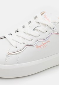 Valkoiset tennarit teksturoidusta nahasta, joissa on aallotettu muotoilu, vaaleanpunainen logo sivulla ja valkoiset tasaiset nauhat. Horisontaaliset uurteet pohjassa.