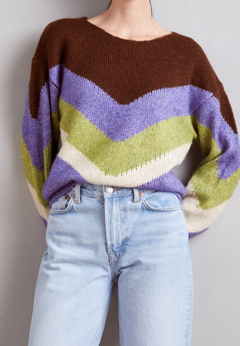 Person wearing a brown, purple, green, and beige chevron-patterned sweater tucked into light blue high-waisted jeans against a neutral background.