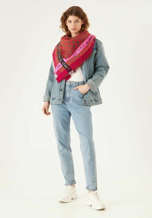 Grande écharpe rose et rouge à motifs avec des bords frangés portée sur une veste en jean, un haut blanc et un jean bleu clair. Des baskets complètent la tenue.