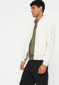 Casaco bomber cinza claro com punhos canelados, fecho de correr à frente e bolsos laterais. Textura suave e design minimalista. Combinado com uma camisa verde.