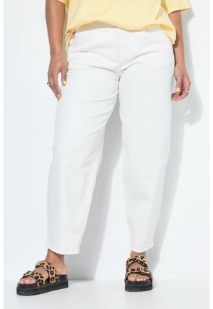 Persona con pantalones blancos, camiseta amarilla, esmalte de uñas amarillo y sandalias de plataforma con estampado de leopardo de pie contra un fondo blanco.