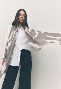 Sciarpa grigia e bianca con motivo a fantasia e accenti scintillanti, realizzata in tessuto morbido, indossata sopra una camicia chiara con bottoni e pantaloni ampi scuri.