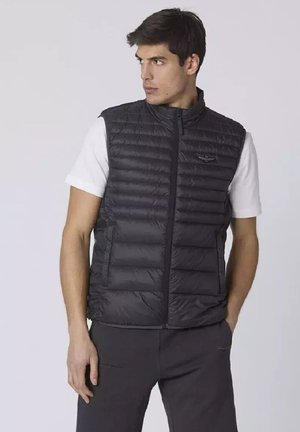 Gilet isolato nero con zip frontale, texture trapuntata e un piccolo logo sul petto, indossato sopra una maglietta bianca e pantaloni grigi.