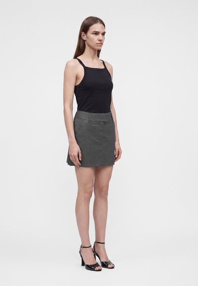 Femme portant un haut noir sans manches, une mini-jupe plissée grise et des sandales noires à talons hauts, debout devant un fond blanc uni.