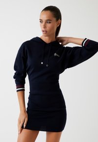 Rochie cu glugă de culoare bleumarin, cu mâneci lungi, având un sistem de strângere cu șnur și accente în dungi. Materialul este neted și ajustat, evidențiind silueta.