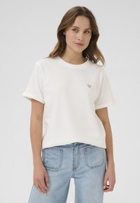 White cotton T-shirt with rolled short sleeves, featuring a small heart design on the chest. Worn with light blue denim jeans.