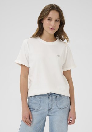 T-shirt en coton blanc à manches courtes rollées, avec un petit motif en forme de cœur sur la poitrine. Porté avec un jean en denim bleu clair.