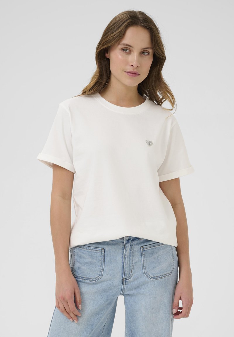 White cotton T-shirt with rolled short sleeves, featuring a small heart design on the chest. Worn with light blue denim jeans.