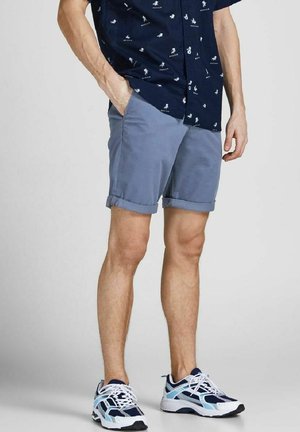 Homme portant une chemise bleue à manches courtes imprimée, un short bleu clair retroussé, et des baskets blanches, bleu marine et bleu clair, debout avec une main dans la poche.