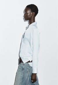 Light blue, button-up shirt with a collar, worn over a denim skirt. The fabric is smooth, and the design is simple with long sleeves.