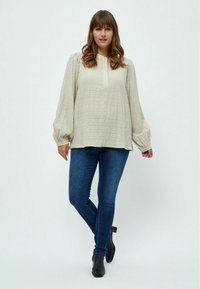 Blusa beige con texture, scollo rotondo, dettagli con bottoni, maniche morbide, abbinata a jeans skinny blu scuro e stivali neri alla caviglia.