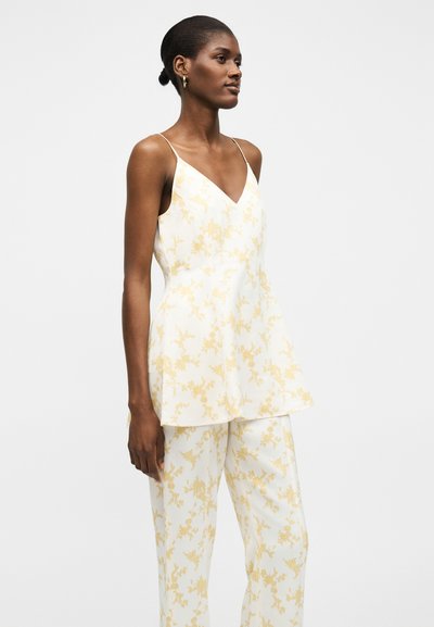 Γυναίκα με λευκό αμάνικο τοπ με V-λαιμόκοψη και παντελόνες με κίτρινο λουλουδάτο σχέδιο, σε χαλαρή στάση μπροστά σε μονόχρωμο φόντο.