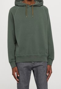 Felpa con cappuccio in misto cotone verde oliva, dal taglio rilassato, con polsini a costine e un piccolo logo sull'orlo. Progettata con cuciture a contrasto.